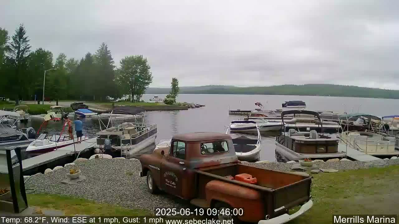 A rusty red pickup truck is parked near a lake marina on an overcast day, with numerous boats docked and people near some of them.