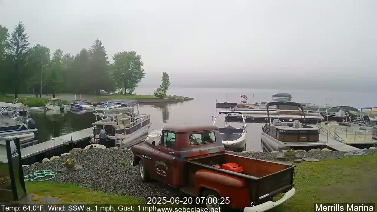 A red vintage truck sits in a foggy marina filled with numerous pontoon boats and other watercraft, near a shoreline with green trees.