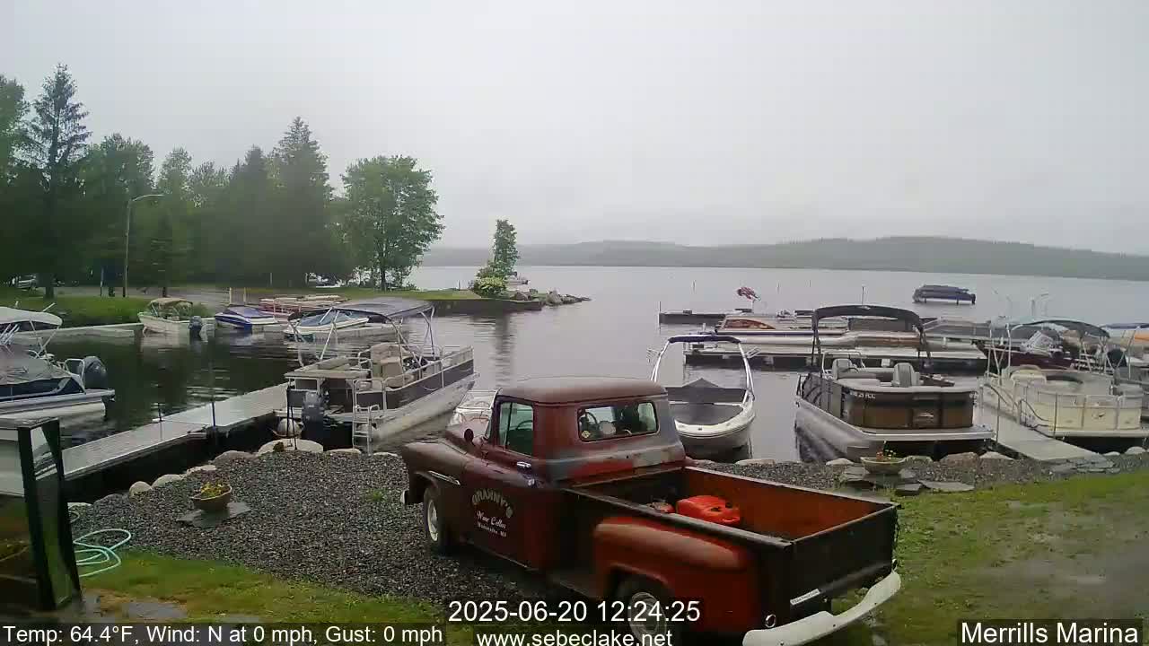 A rusty red pickup truck sits in a marina on an overcast day, surrounded by numerous boats docked along a shoreline with green trees in the background.