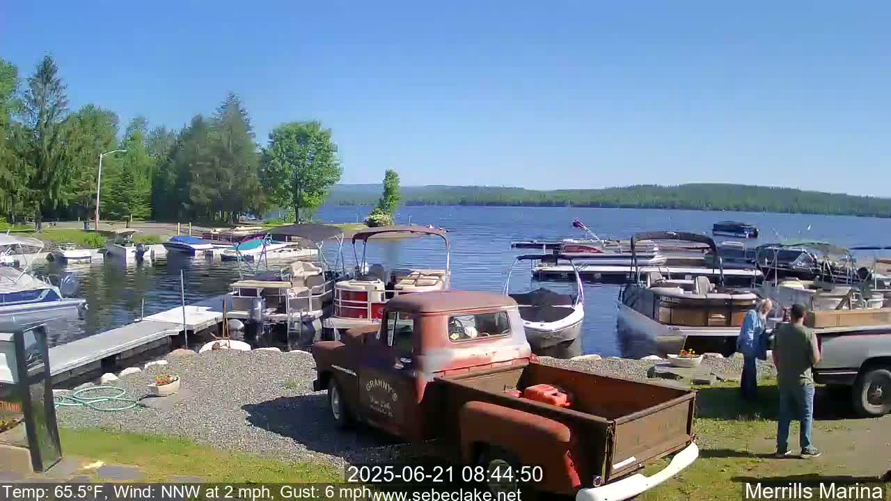 A sunny day at a marina shows numerous boats docked along a shoreline with a rusty red truck parked nearby, and people standing near the boats.