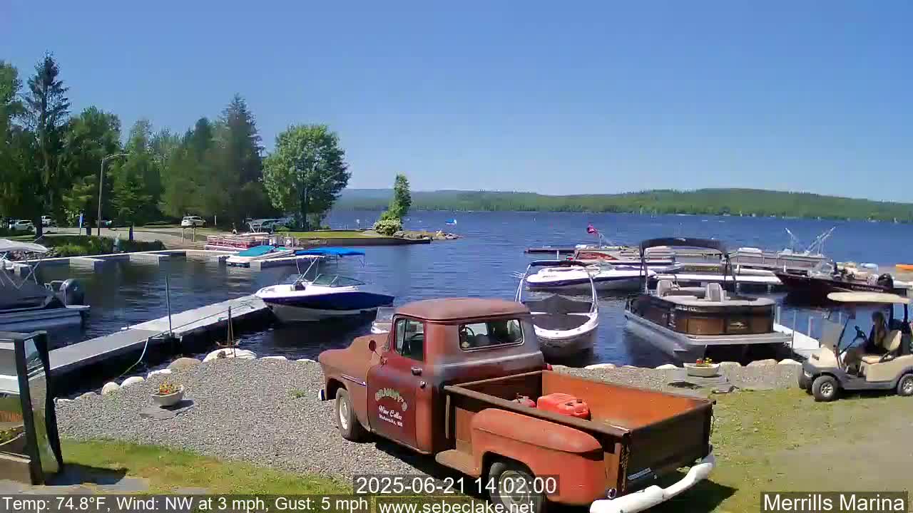 A sunny day at a marina shows numerous boats docked, a rusty red pickup truck, and a golf cart parked on a gravel lot next to a calm lake surrounded by green trees.
