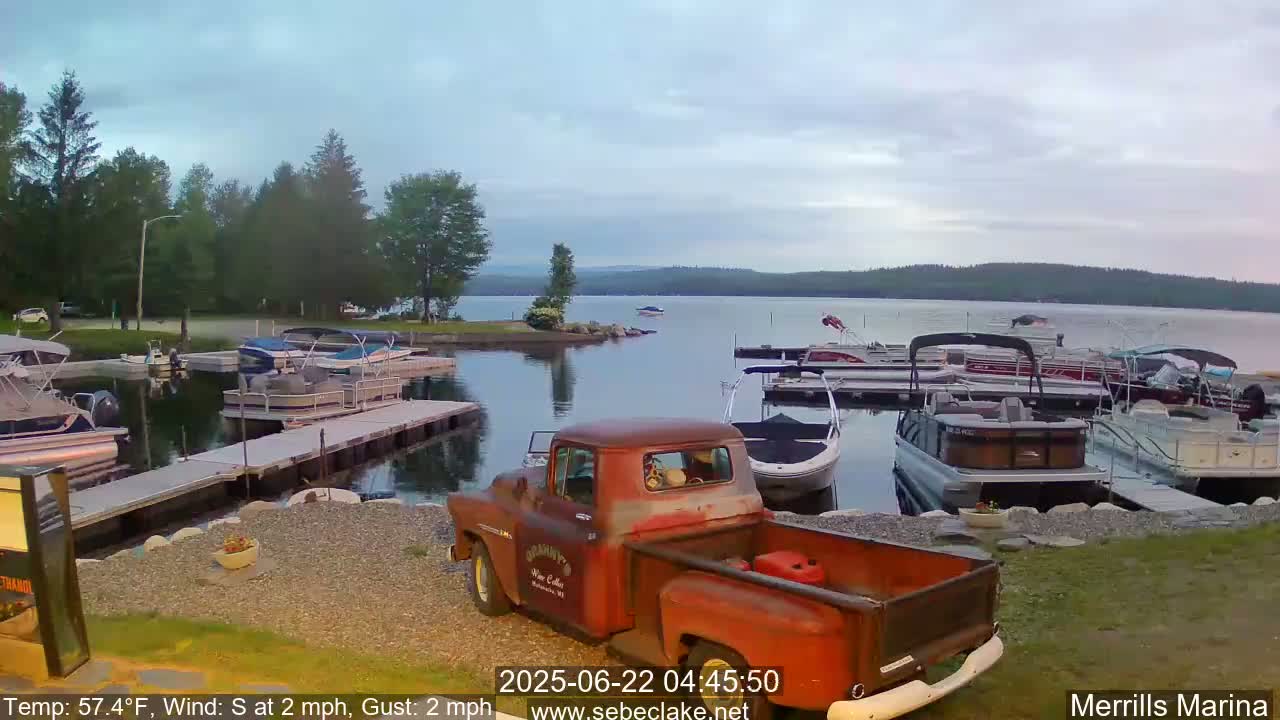 A rusty red pickup truck sits in a marina at dawn, numerous pontoon boats are docked along a calm lake under a partly cloudy sky.