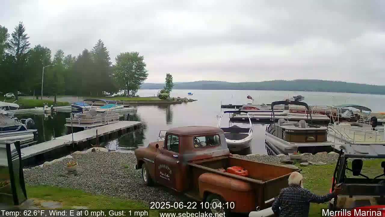 A rusty pickup truck is parked near a marina with numerous boats docked, all under an overcast sky.
