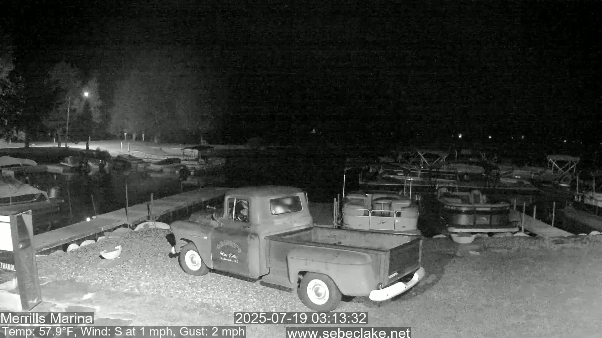 A nighttime, low-light grayscale image shows a vintage pickup truck parked in a marina with numerous boats docked nearby under a dark, clear sky.