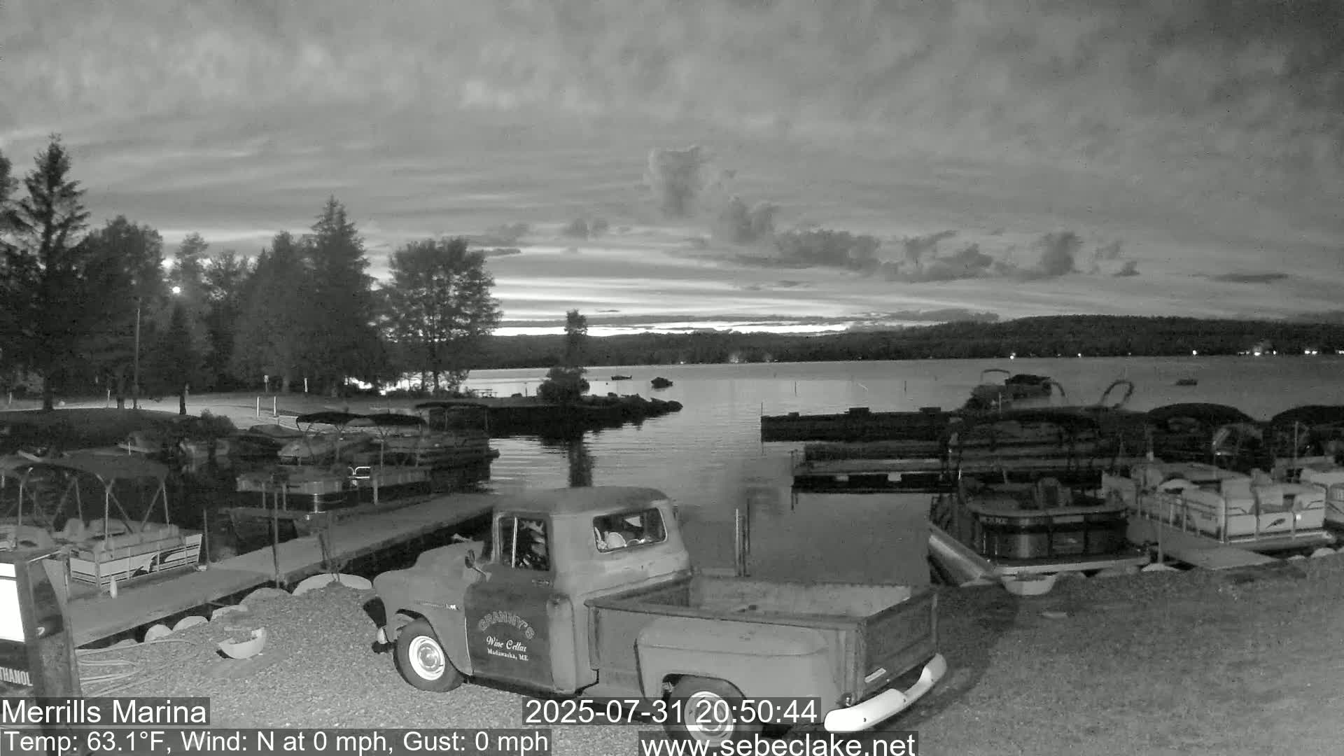 A mostly cloudy twilight scene shows a lake marina with several pontoon boats docked, and a vintage pickup truck parked nearby.