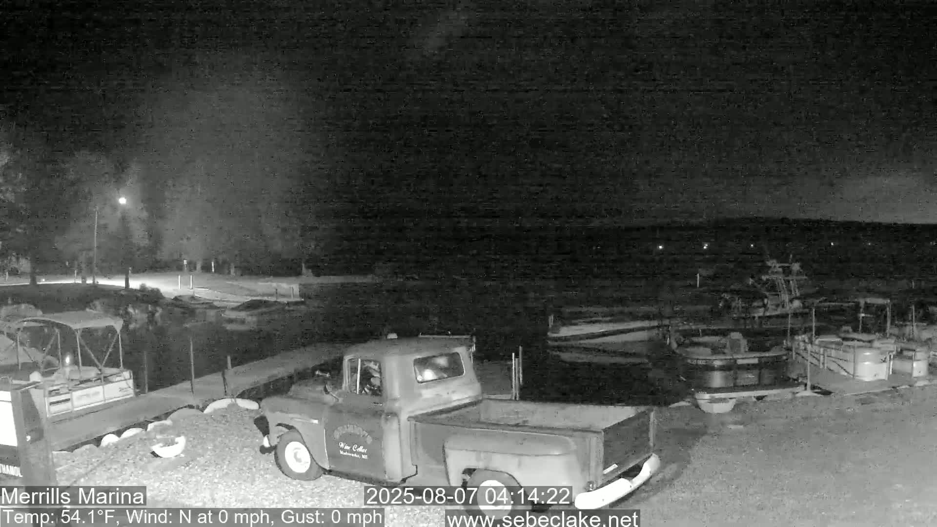 A nighttime view of a marina shows several boats docked, a pickup truck parked nearby, and calm conditions with no wind.