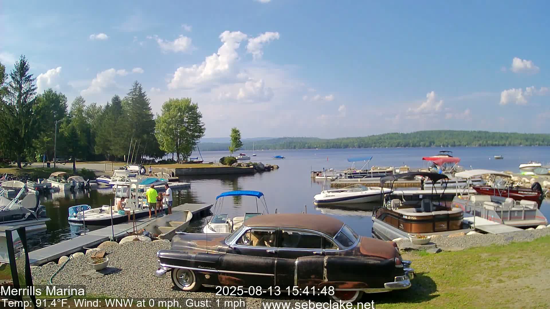 A classic car sits near a marina filled with boats on a sunny day with light clouds and a gentle wind.