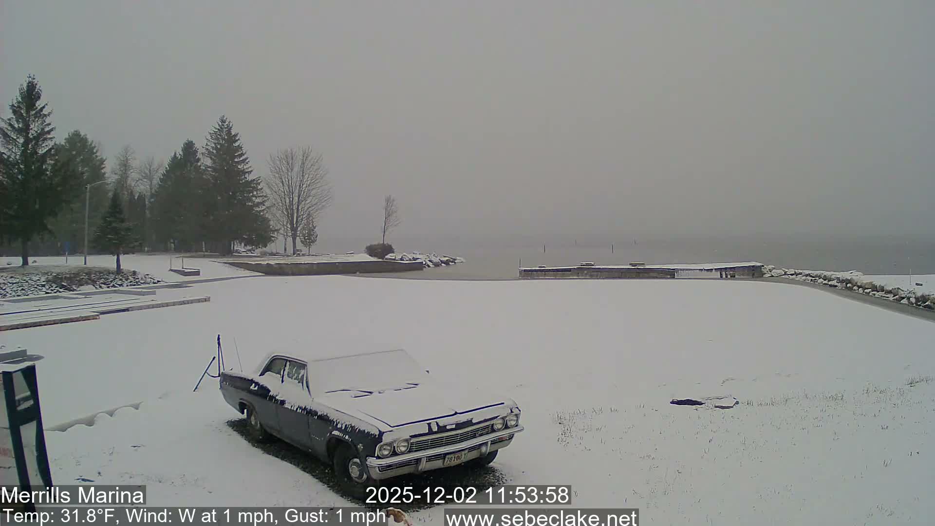 A snow-covered dark classic car is parked at a marina with docks extending into a hazy lake, all under a grey and overcast winter sky.