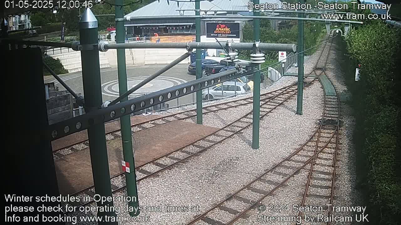 A high-angle view shows a tram station's tracks curving into a tunnel, with several cars parked nearby in a sunny parking lot.