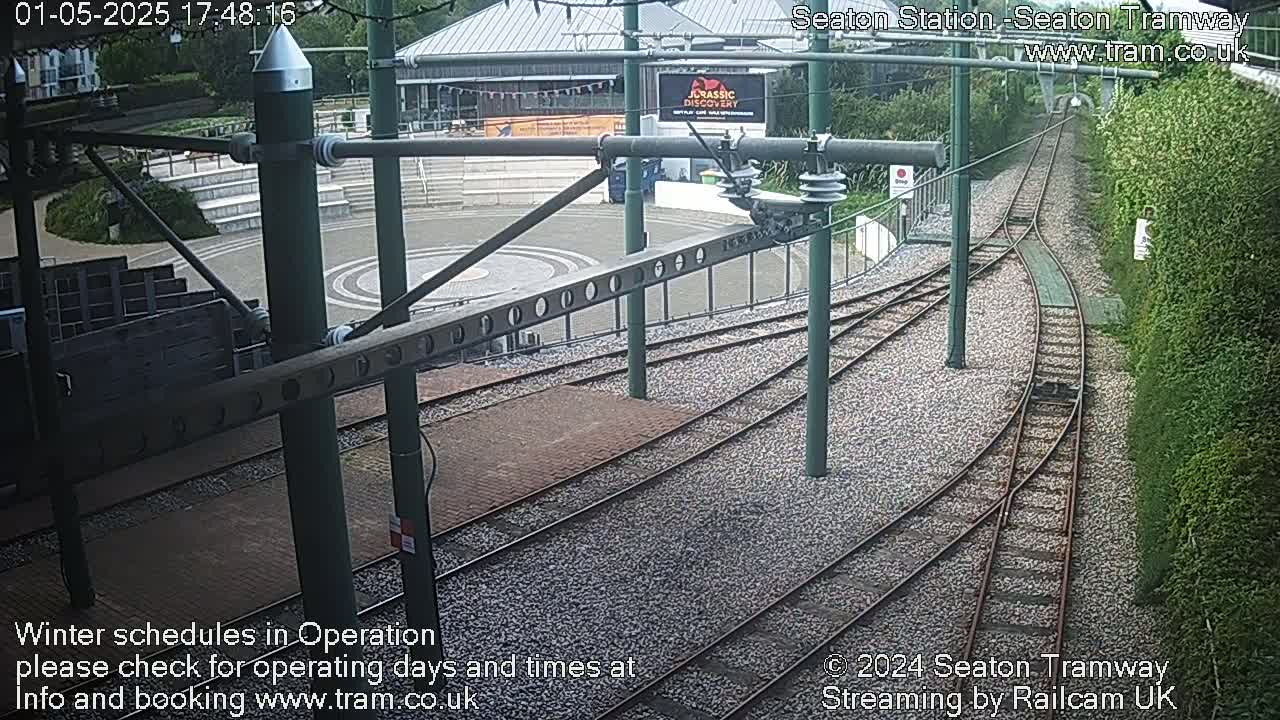 Tram tracks curve around a station platform, next to a building and greenery, in overcast conditions.