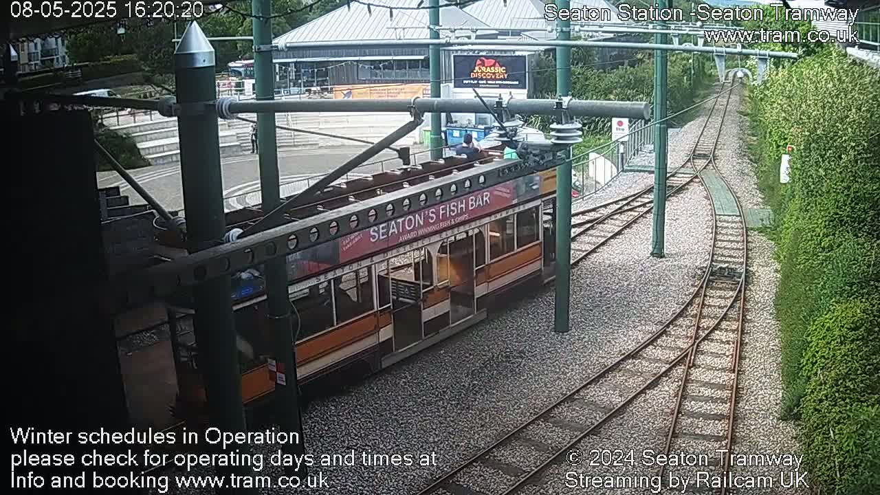 An orange and brown tram is at a station, with a split rail track leading away from it.