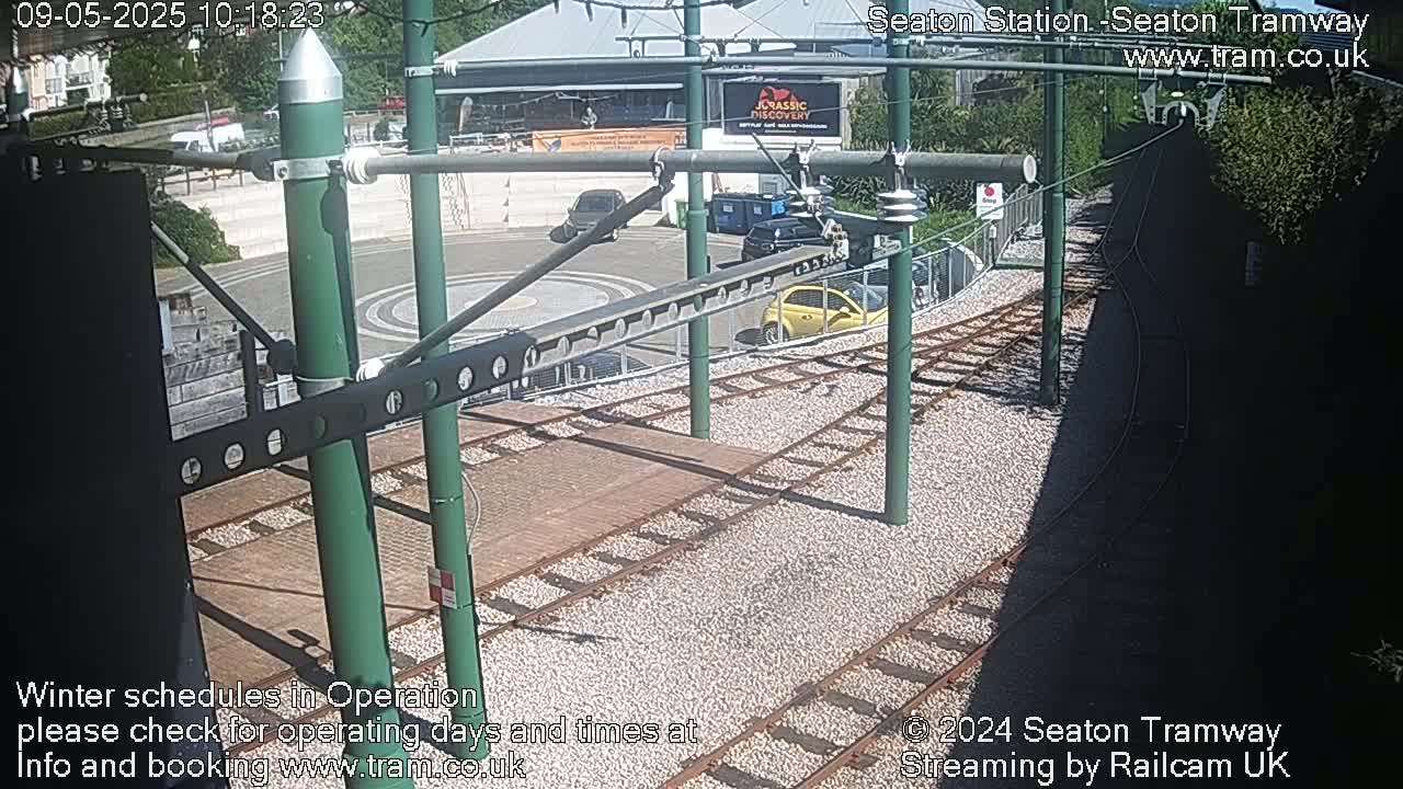 A sunny view of tram tracks and a station platform, with several cars parked nearby.