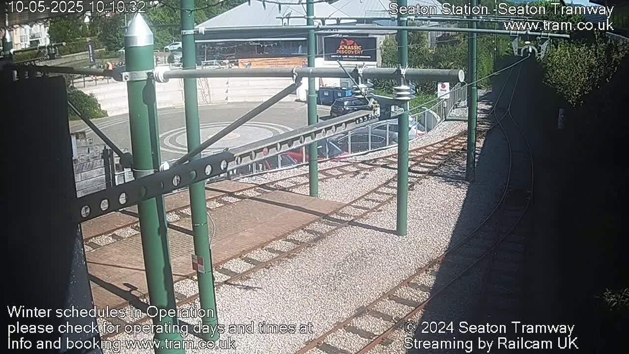 A sunny overhead view of tram tracks and a station area with a few parked cars and a building in the background.
