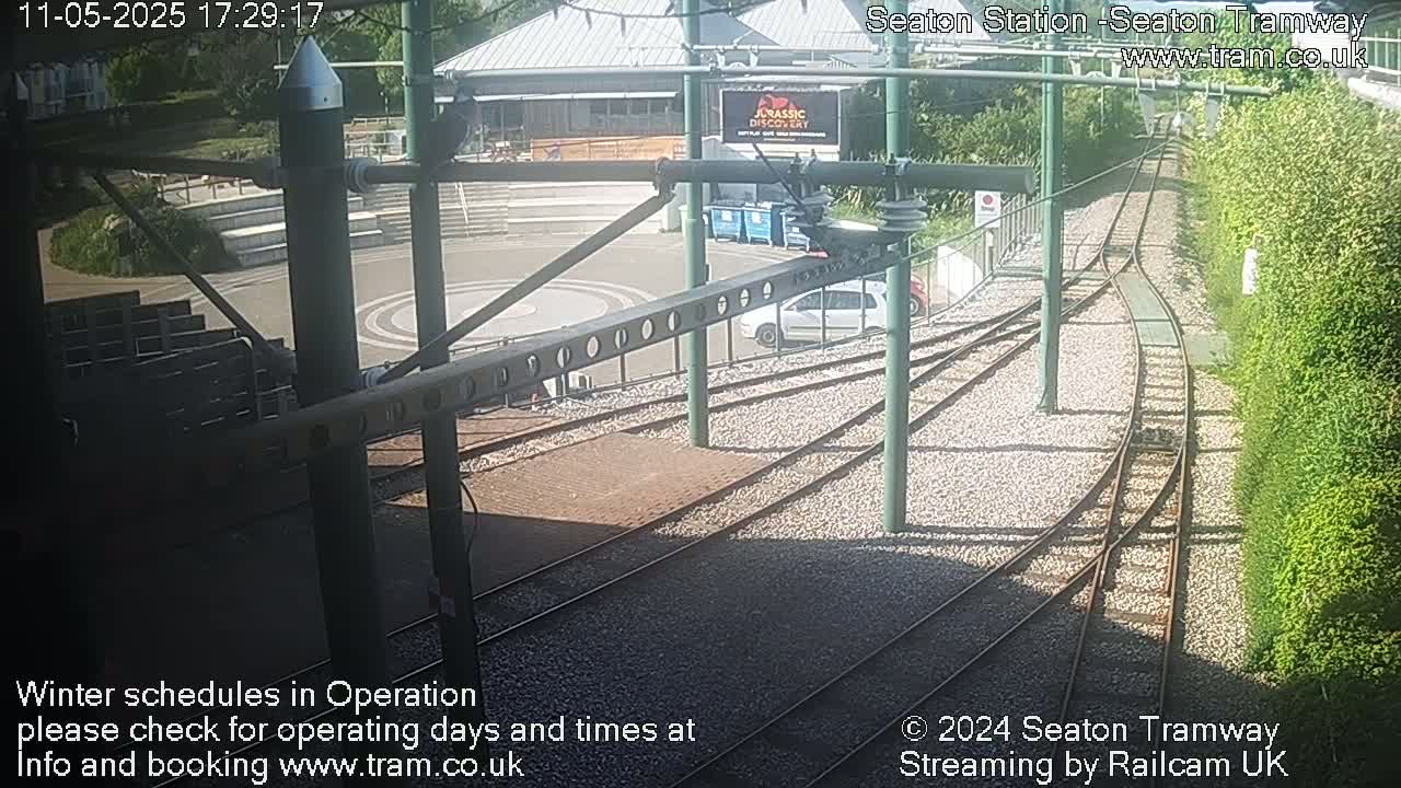 A sunny view of train tracks curving around a station platform, with a car parked nearby and lush greenery bordering the scene.