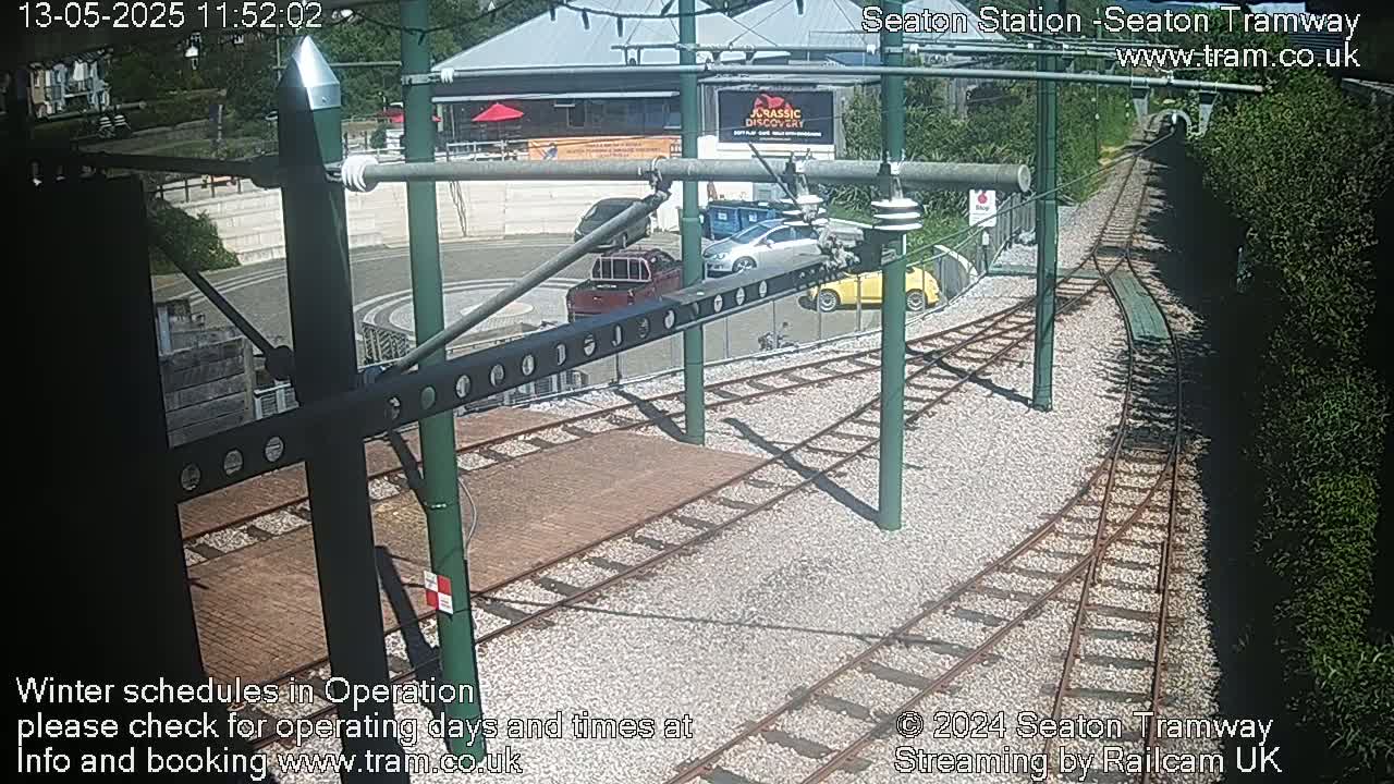 A sunny view of a tram station's track area with several parked cars, showing a convergence of railway lines and surrounding greenery.