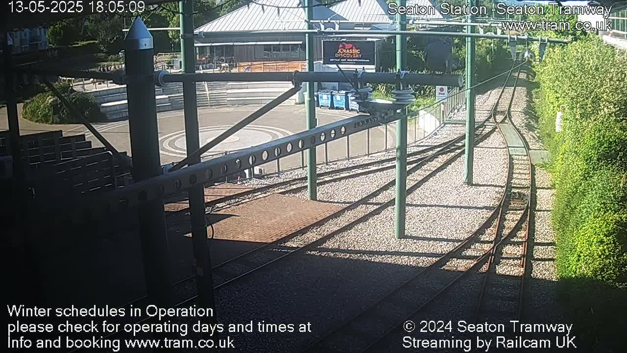 A sunny outdoor view shows a tram station with train tracks curving away from a platform and into a green hedge.