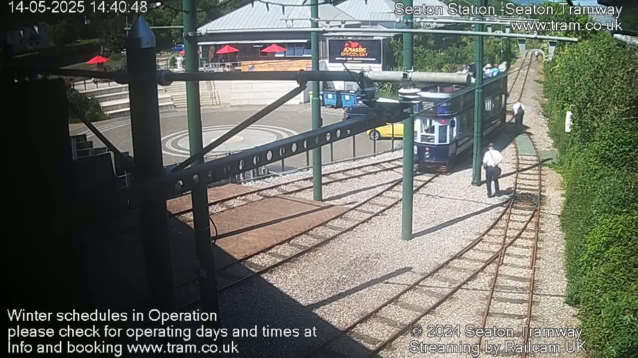 A tram is stopped at a station on sunny day, with a person walking nearby, and another person standing near a building in the background.