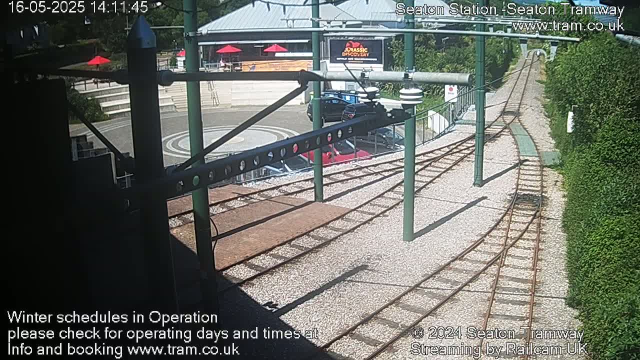 A sunny view of a tram station with tracks curving away, several parked cars, and a building in the background.