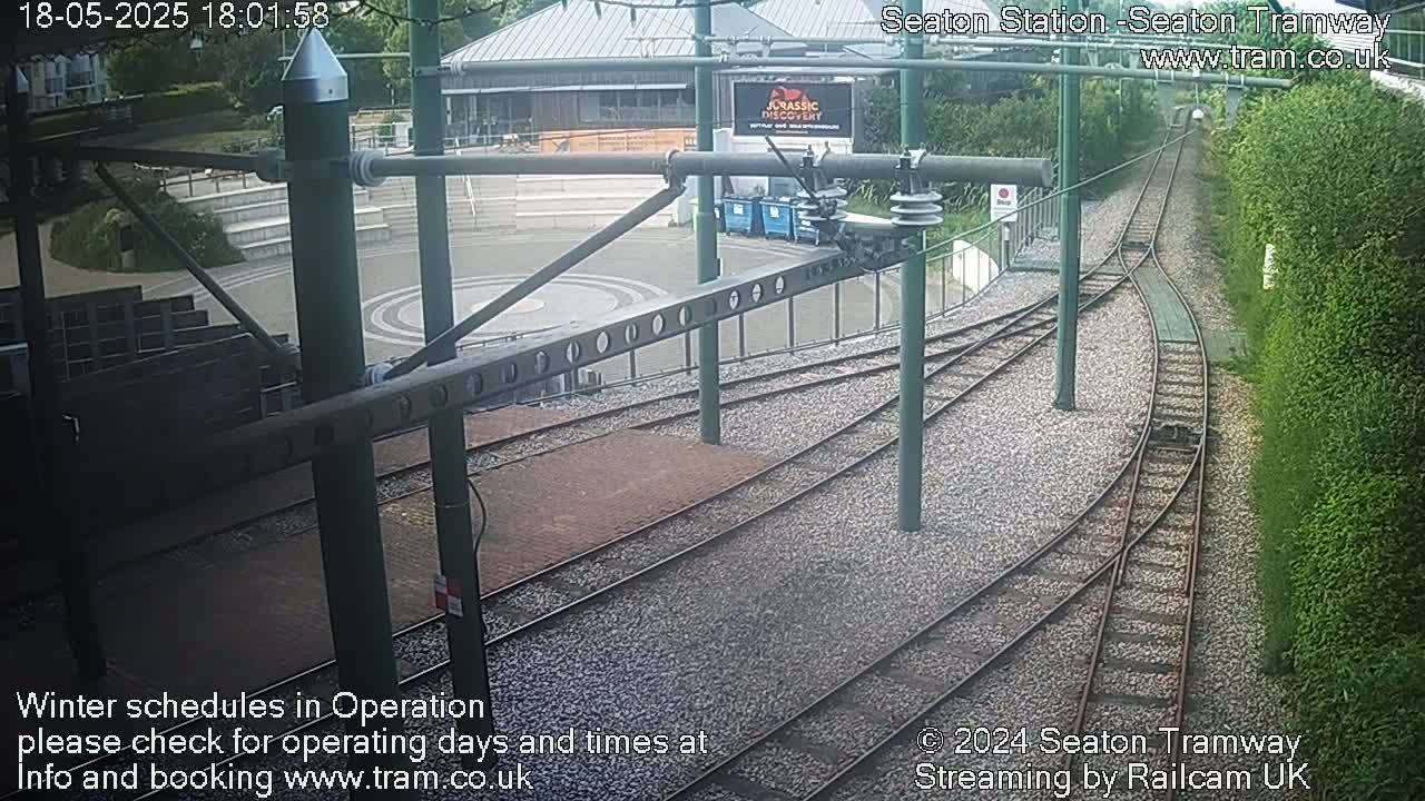 A high-angle view of a tram station's tracks and platform, with a building and greenery in the background under overcast skies.