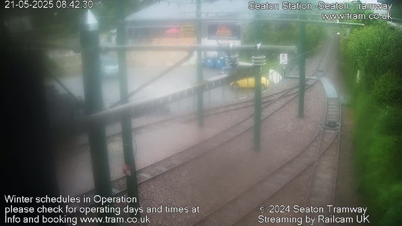 A rainy view of a tram station with train tracks and a few vehicles visible.