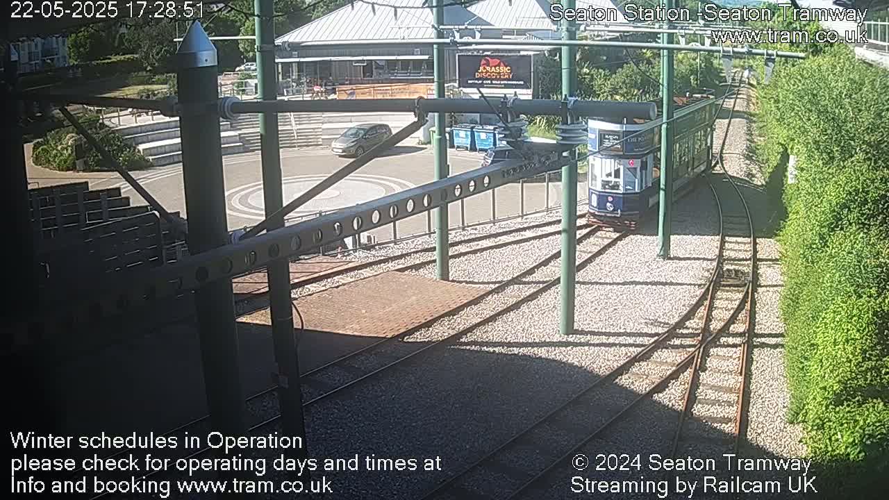 A tram approaches a station on a sunny day, passing alongside a car and a building.