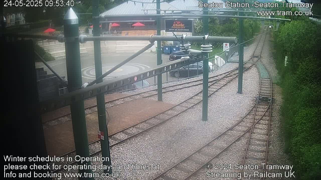A tram station's overhead view shows a turning section of railway track, cars parked nearby, and a building with outdoor seating in the background under an overcast sky.