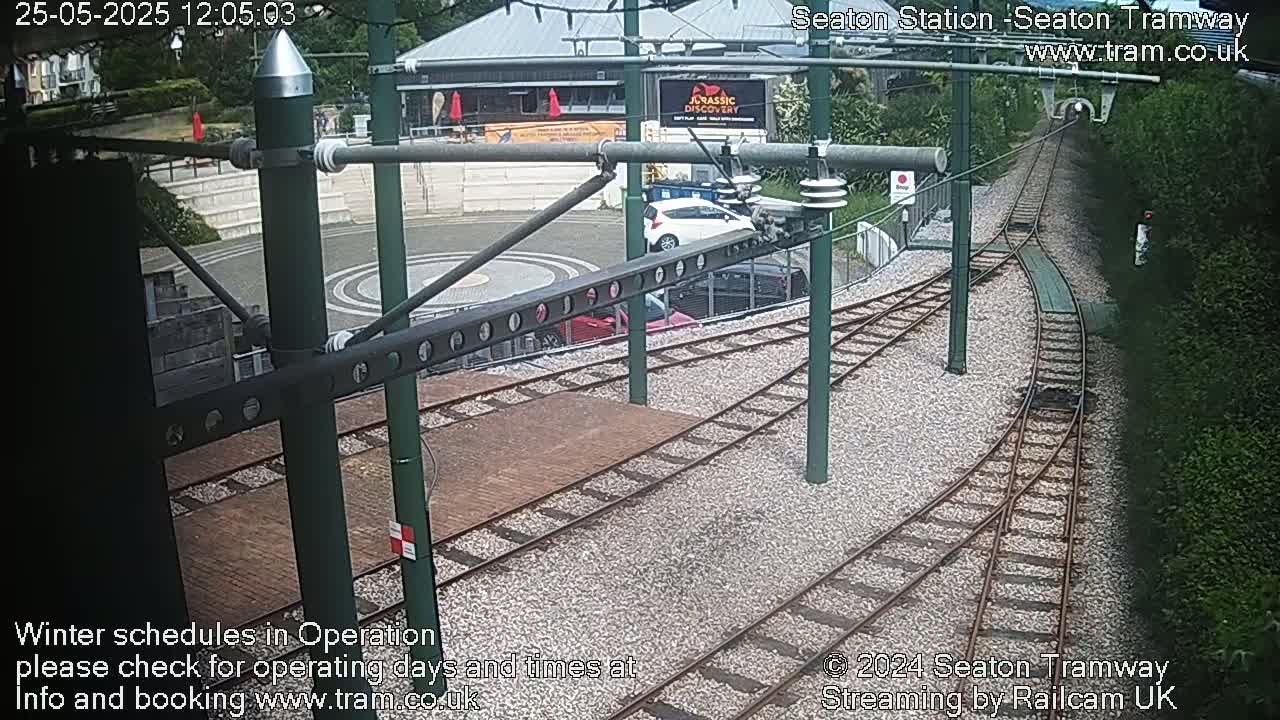 A high-angle view of a tram station with train tracks branching off, several cars parked nearby, and lush green hedges under a partly sunny sky.