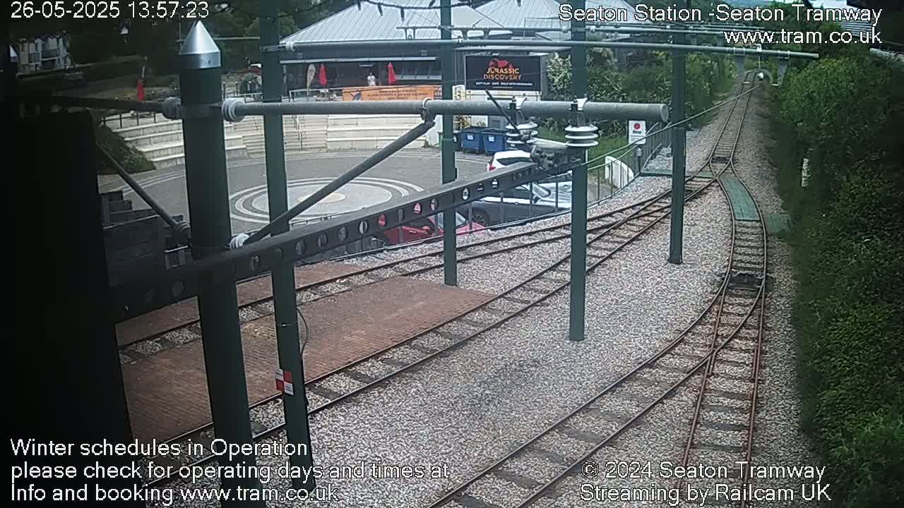 Overhead tram tracks curve away from a station platform with several parked cars visible; the weather appears to be clear and sunny.