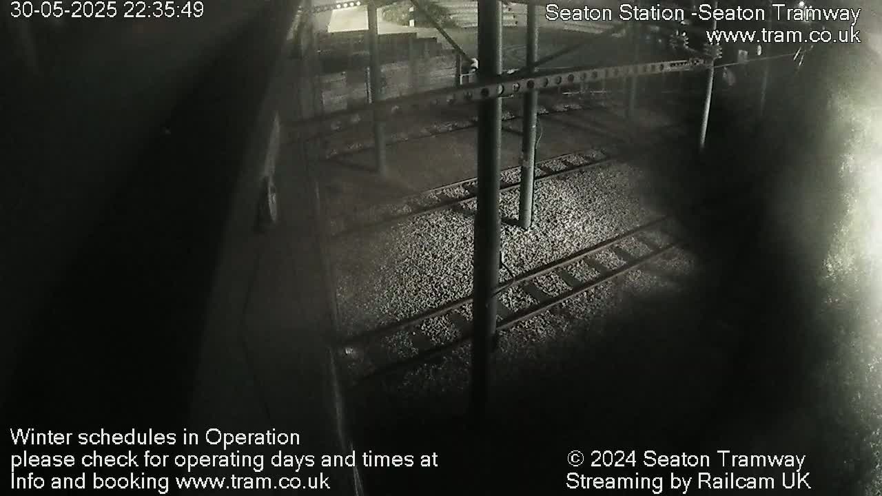 A nighttime overhead view of train tracks covered in light-colored gravel under a metal structure.