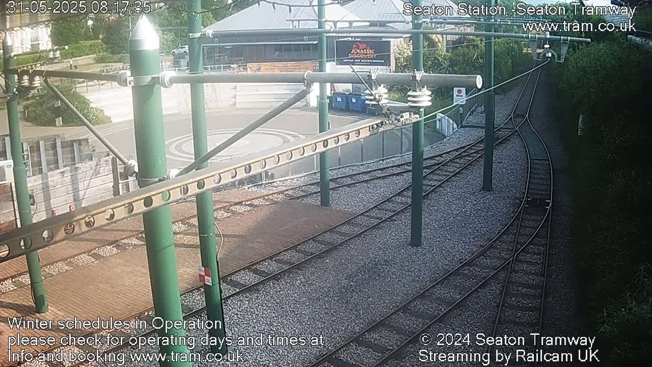 Overhead tram lines and tracks curve around a station platform on a sunny day.