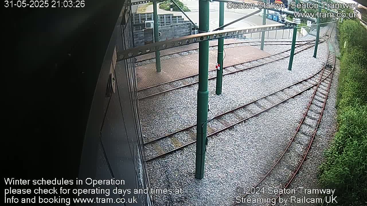 A high-angle view shows several train tracks curving away from a station platform, surrounded by gravel and vegetation.