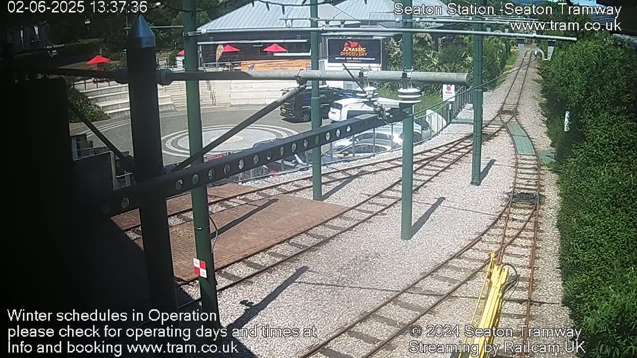 A sunny view of a tram station with train tracks, a turning loop, several parked cars, and surrounding greenery.