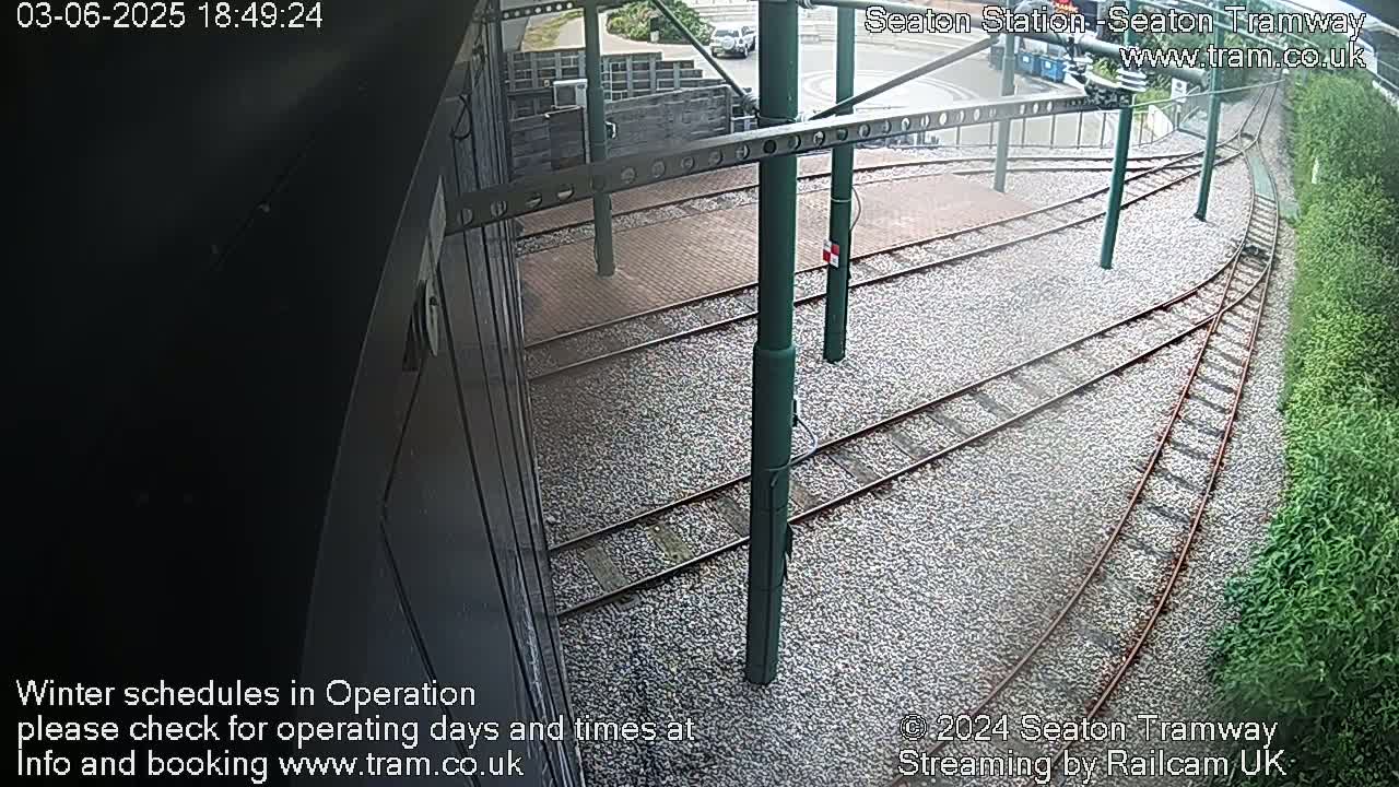 A high-angle view shows tram tracks curving away from a station platform, set in a gravel bed next to a hedgerow.
