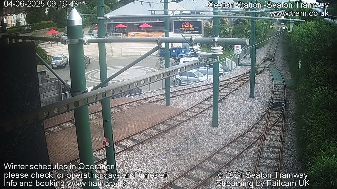 Overhead tram tracks curve through a station area with several parked cars, buildings, and lush green vegetation visible under an overcast sky.