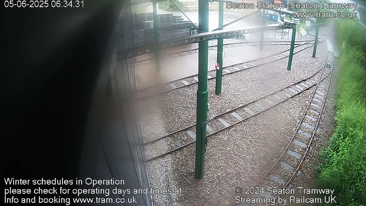 Tram tracks curve through a gravel area beside vegetation on a wet day.