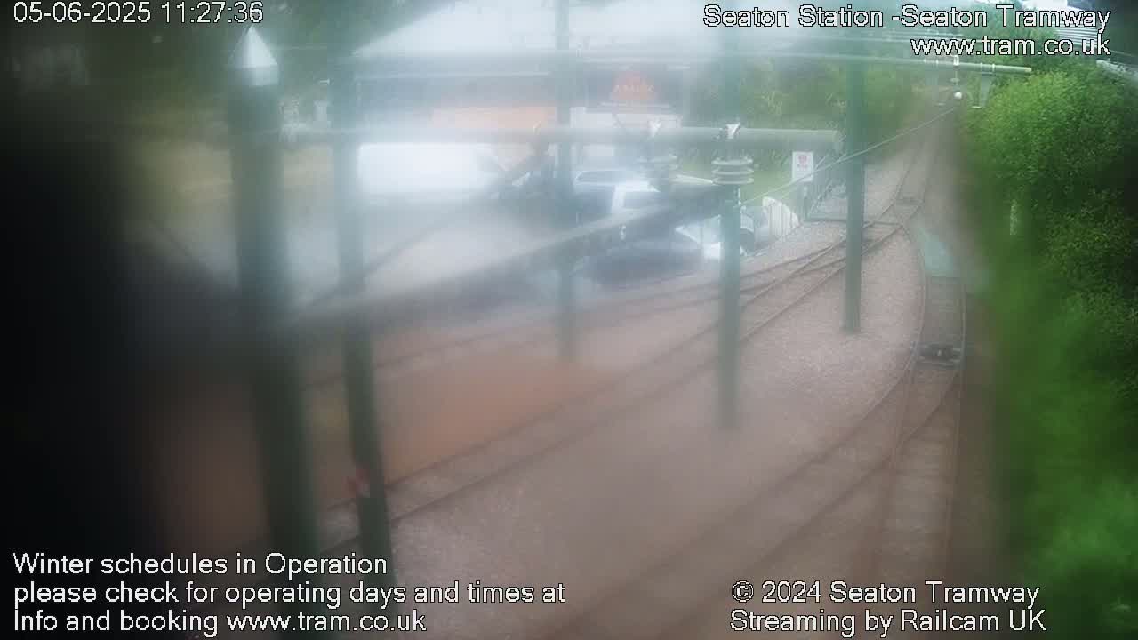 A hazy, rainy view of tram tracks curving through a station area, with cars parked nearby and green vegetation visible.