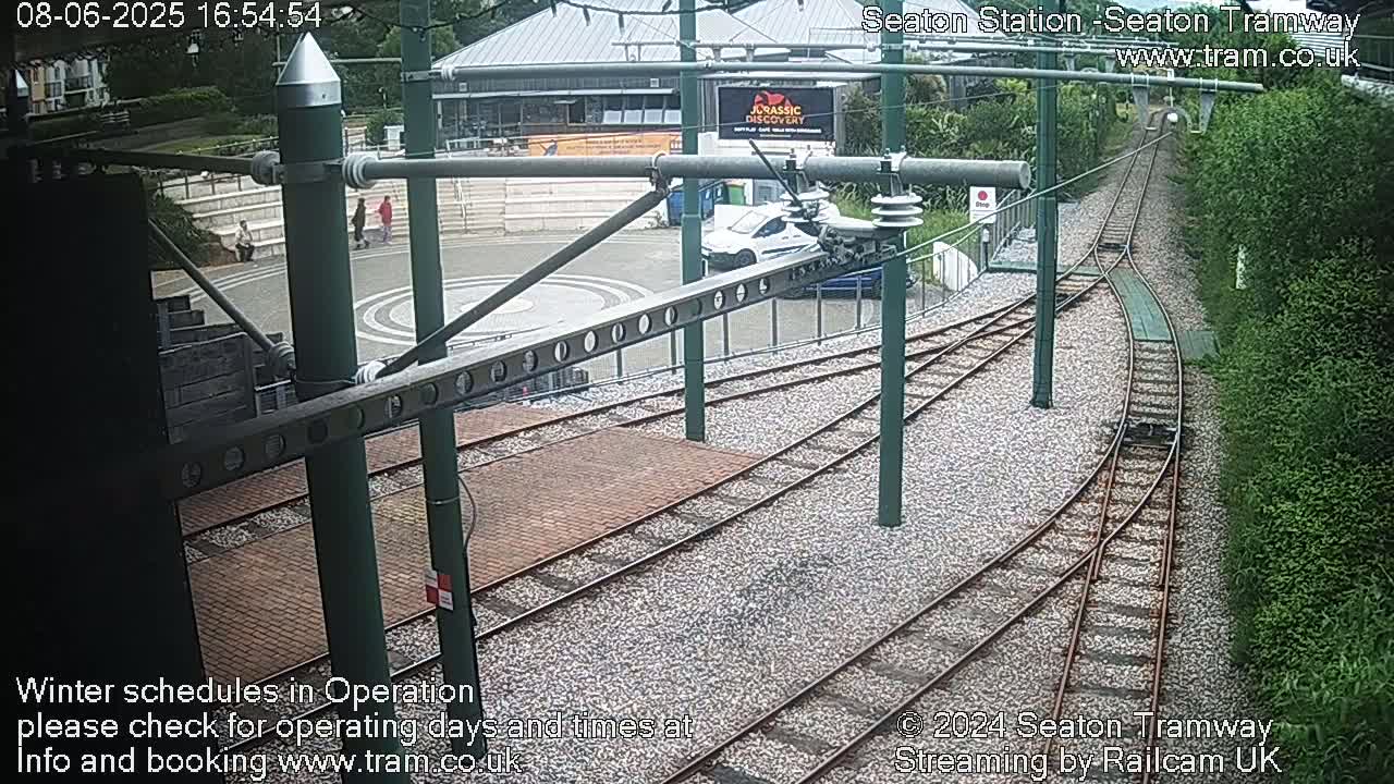 A high-angle view shows a tram station with railway tracks curving away from a brick paved area, a car parked nearby, and some people walking; the weather appears overcast.