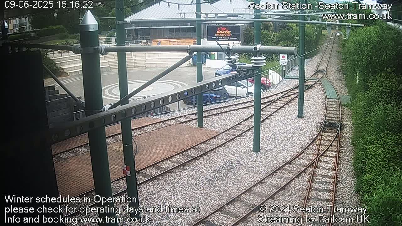 A high-angle view shows a tram station with several cars parked nearby, tram tracks curving away from the station, and lush green vegetation bordering the tracks.