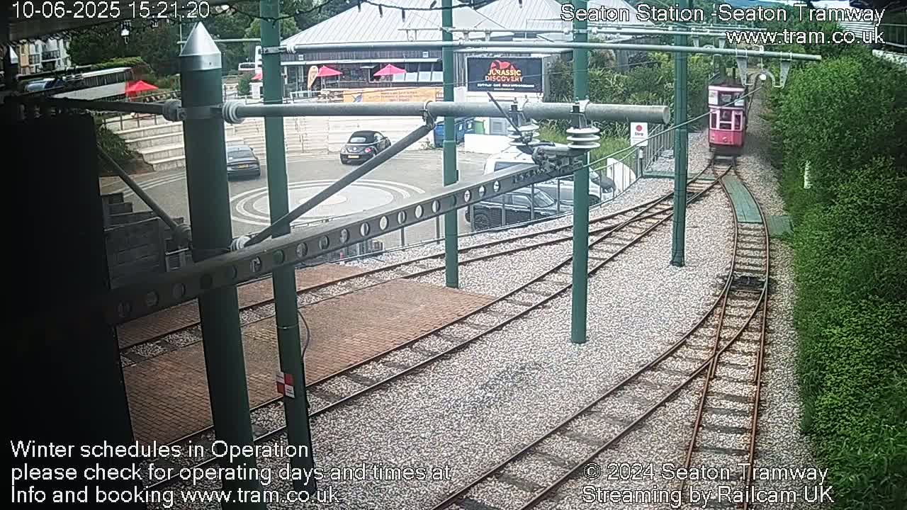 A pink tram travels along a railway track that curves to the right, passing by a station area with cars parked and a building in the background, on a sunny day.