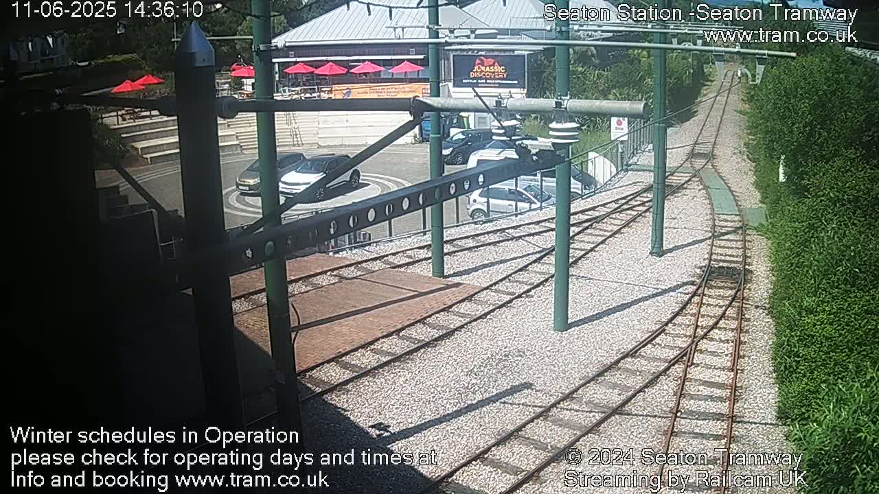 A sunny outdoor scene shows tram tracks curving around a station platform with several cars parked nearby, and a building with red umbrellas visible in the background.