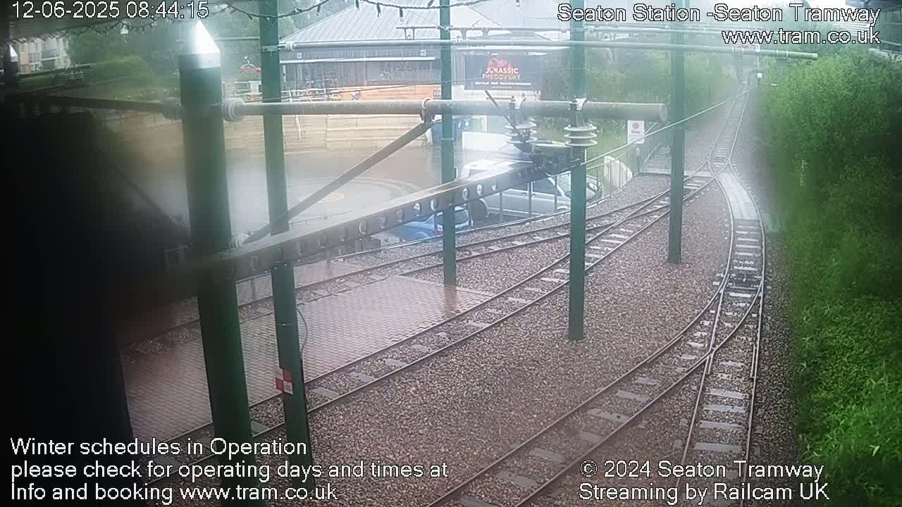 A rain-soaked tram station with tracks branching off, showing some vehicles parked nearby.