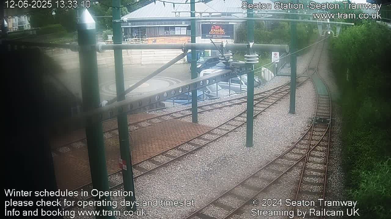 Tram tracks curve through a gravel area near a building, with cars parked nearby, under an overcast sky.
