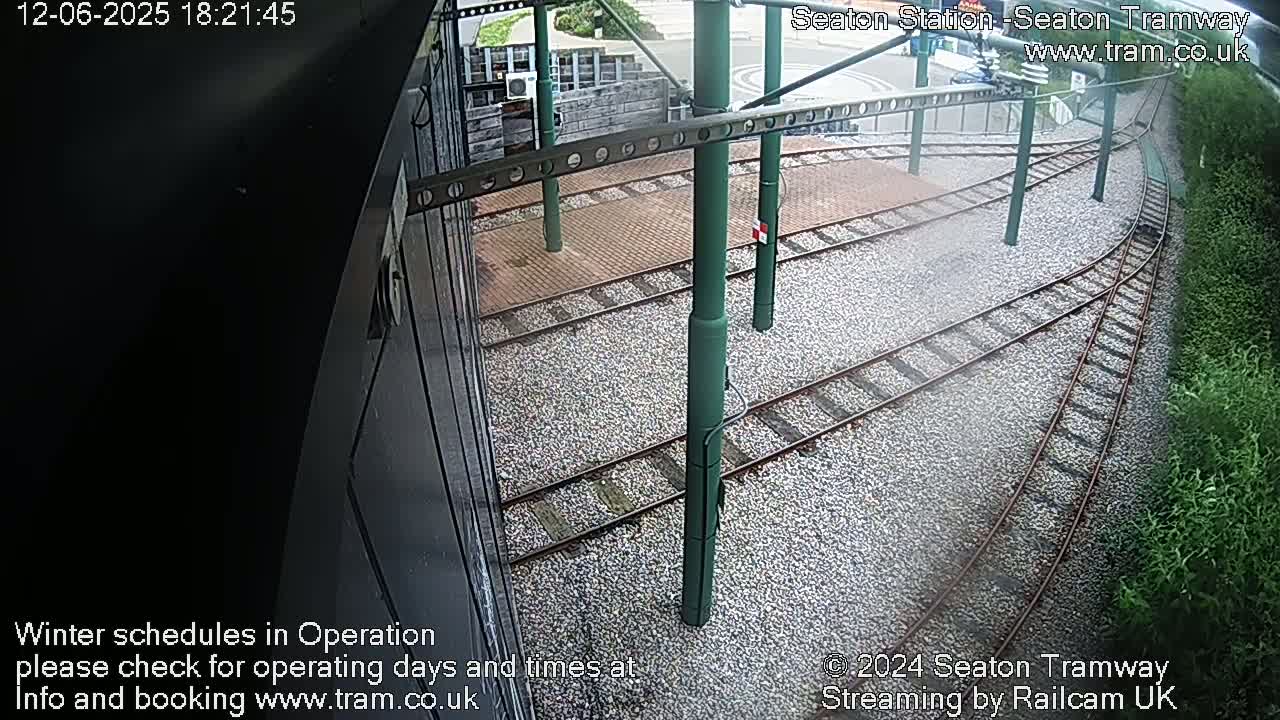 A high-angle view shows train tracks curving through a gravel area next to some greenery, under overcast conditions.