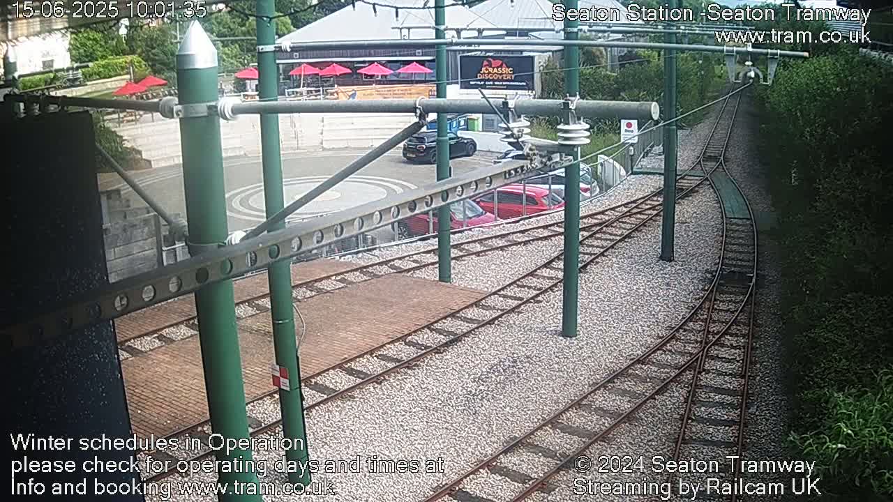 Tram tracks curve through a gravel area near a building with red umbrellas, some parked cars, and green vegetation.