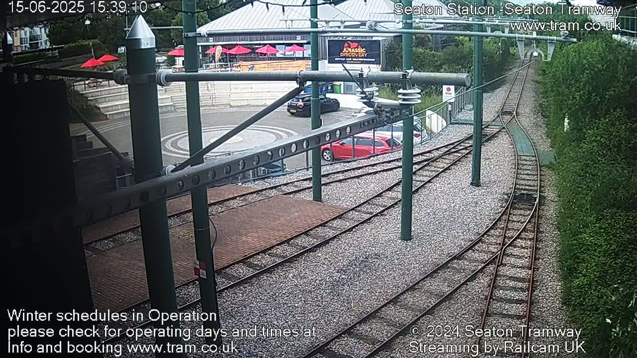 Tram tracks curve through a station area with parked cars and a building visible in the background under a partly sunny sky.