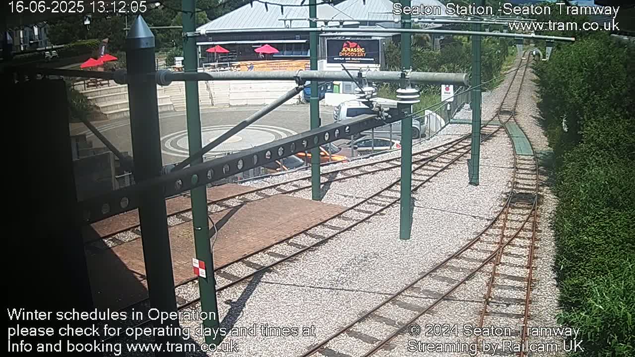 A sunny view of a tram station with train tracks curving away from a paved area, a building in the background, and green hedges on either side.