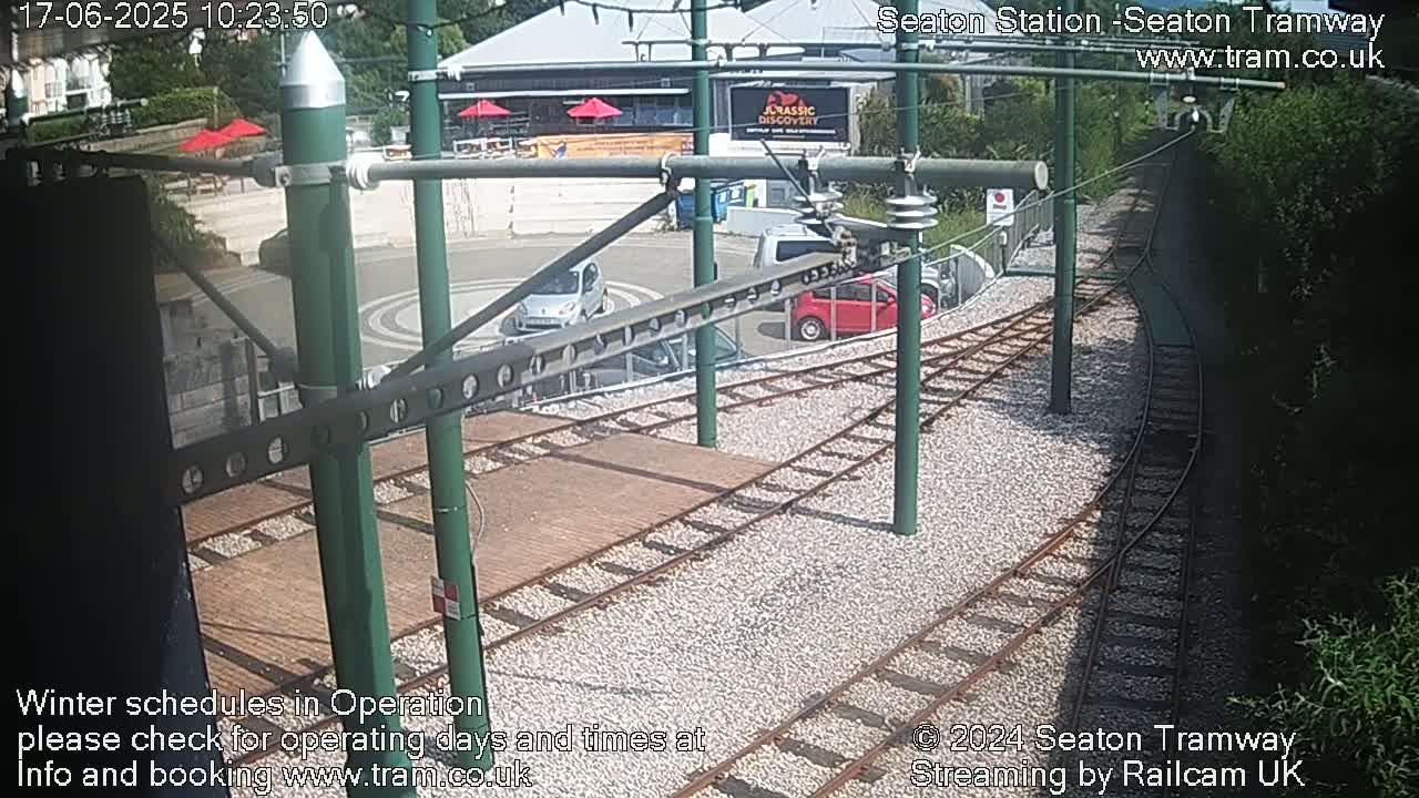 A sunny view of a tram station with several cars parked nearby and train tracks leading away.