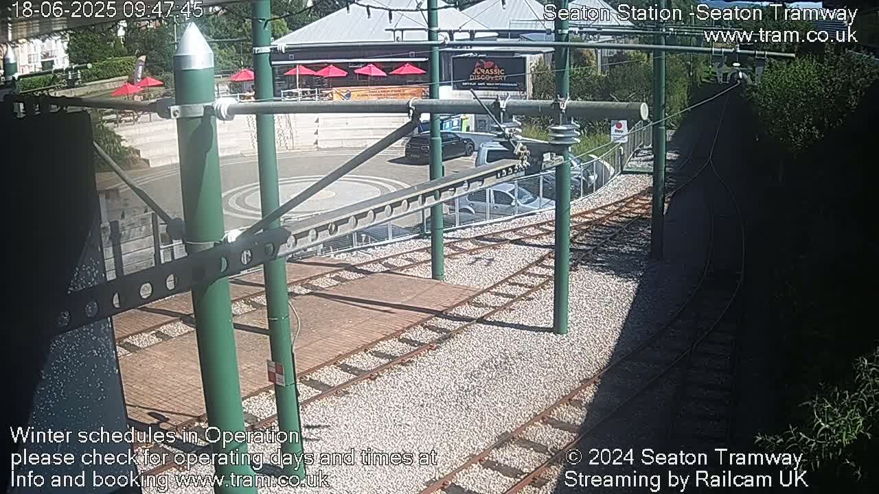 Tram tracks curve through a sunny outdoor plaza with parked cars and a building with red umbrellas.