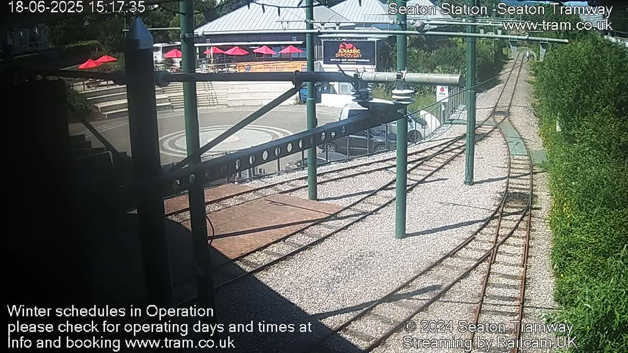 A sunny outdoor view of tram tracks curving through a gravel area next to a building with red umbrellas and lush green vegetation.