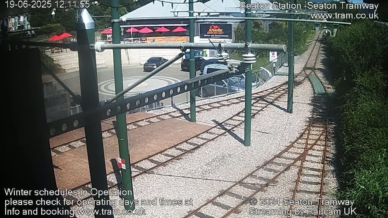 A sunny outdoor scene shows a tram station with several cars parked nearby and train tracks leading away from the station, bordered by hedges.